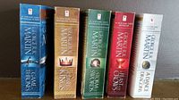 Five paperback books standing upright showing spines: Game of Thrones (blue), Clash of Kings (brown), Storm of Swords (green), Feast for Crows (dark red), and A Dance with Dragons (white). Each spine displays title, author, and publication branding.