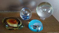 Four glass art paperweights displayed together: large clear bubble paperweight, clear blue swirl paperweight, amber swirl diamond shape, blue half circle butterfly paperweight.