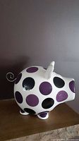 Side view of ceramic piggy bank showing purple and black polka dots, metal spiral tail, and overall shape.