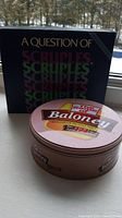 Photo shows both The Game of Baloney tin can with vintage label and A Question of Scruples Second Edition box.