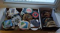 Image showing multiple stacks of square and circular vintage bar coasters in three boxes on a windowsill, assorted designs and brands visible.