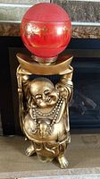 Full view of the gold-colored heavy duty plastic Buddha statue holding a red glass ball light above its head, placed in front of a fireplace.