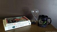 Two paperback books stacked on a wooden surface alongside a glass hourglass sand timer and a black ceramic mug with Wicked Witch design and quote
