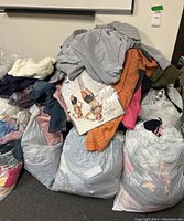 Large bags of mixed women's clothing piled together, showing a grey sweatshirt, orange checked shirt, and various other assorted fabric colors.