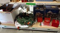 Photo showing a boxed Christmas village set labeled 'Workbench' with two small snowy buildings, a holiday wreath with assorted decoration, a red mug with treat bags, and a large shipping box at the bottom.