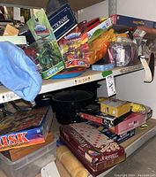 Shelf showing multiple board games including Risk, Scrabble, and Pictionary, along with toys and figurines in packaging stacked and stored in a cluttered manner.