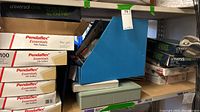 Shelf with stacked boxes of Pendaflex Essentials file folders and Universal One binders, a blue magazine file holder, and a green plastic container below.
