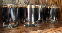 Nine black lowball glasses with gold trim arranged in front of a wood background.