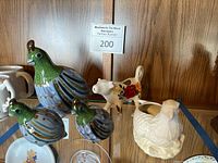 Overview of momma quail and baby quail figurines next to cow creamer and eagle pot on glass shelf.