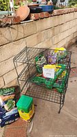 Photo showing black metal wire garden stand with bags of plant food and garden soil under and inside it along with small plant pots on the brick wall above