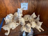 Front view of all seven white Lenox cat figurines displayed on glass shelf, showing variety in poses and decorations including bows and jeweled collars.