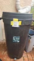 Full frontal view of black cylindrical rain barrel with yellow tag and Ivy brand label near middle, positioned outside on brick surface beneath a white downspout.