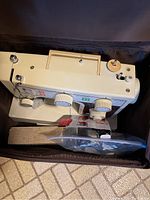 White portable sewing machine in vertical orientation showing knobs, spool pin, handle, and attached cords.
