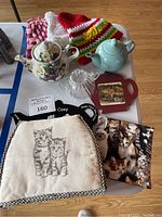Overview photo showing all three teapots, multiple tea cozys, and a red trivet with a rural scene, arranged on a white table.