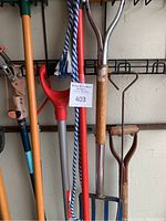 Various garden tools hanging on wall rack including shovels, pitchfork, rakes, high branch saws, and long-handled implements.