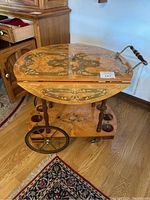 Full view of round vintage wooden tea cart with floral and scroll designs, hinged top, and wooden wheels.