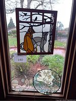 Craftsman style stained glass panel showing two cats and birds framed in dark wood, hanging by chain in a window with garden view