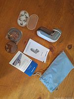 Overhead view of white and gray fabric shaver with digital display, replacement shaving heads, protective covers, instruction manuals, and blue fabric storage pouch on wood surface.