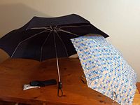 Two umbrellas side by side on a wooden table: one black compact with sleeve, one blue patterned full-size umbrella open
