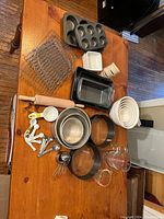 Photo showing muffin tin, cooling racks, rectangular pans, mixing bowls, rolling pin, measuring spoons, and small containers on wooden table.