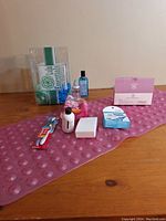 Photo showing assorted bathroom items arranged on wooden surface including pink bath tub grip mat, razors, toothbrush, scrubbies, soap bottles, mouthwash, and cotton pads.