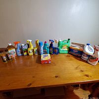 Full display of cleaning products lined up on wooden table showing wide variety of liquid detergents, sprays, bleach, and soaps.