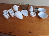 Image showing a set of white ceramic serving pieces including cups, teapot, creamer, bowls, a metal creamer, and wire basket on wooden table.