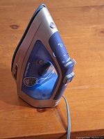 Blue and silver Hamilton Beach iron standing upright showing the side profile, highlighting the retractable cord button and splash symbols for water and steam.