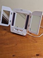Front view of the white tri-fold makeup mirror with side lights, control knobs, and unplugged power cord, showing all three mirrored panels on a wooden surface.