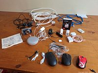 Lot contents on wooden surface showing Fujifilm camera, Google Nest Mini, cables, adapters, and computer mice.