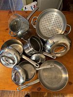 Image showing the assortment of stainless steel pots, pans, lids and strainer baskets arranged on a wooden table.