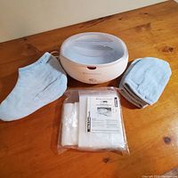 Photo showing the Remington paraffin wax heating system unit, light blue gloves, foot covers, and packaged disposable liners on wooden surface.