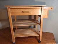 Front view of wooden kitchen cart showing drawer, two slatted shelves, wooden towel bar on side, and caster wheels.