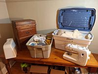 Photo of wooden sewing cabinet with white protective case, plastic tubs with fabric and sewing accessories, and Janome PRL 18 sewing machine on tabletop.