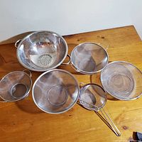 Top view of six kitchen strainers and colander arranged on wood table showing sizes and handle types