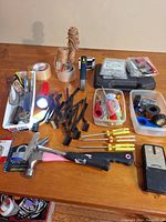 Overview of all hand tools and accessories on wooden table including hammers, screwdrivers, adhesive tapes, flashlight, wire brushes, twine, stud finder, and small hardware items.