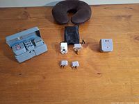 Photo showing three travel voltage adaptors, a brown neck pillow, and a grey plastic case containing one adaptor.