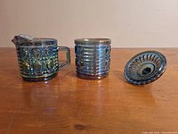 Blue carnival glass creamer, covered sugar bowl, and lid shown from the side on a wooden surface