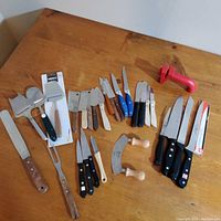 Photo showing various kitchen cutting tools including knives of different sizes, cheese knives, a mezzaluna chopper, and a red knife sharpener on wooden handles.