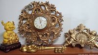 1960s gold resin Rococo style wall clock, large gold decorative key, gold gilded cherub wall hanging, and golden coin cow figurine on wood base arranged on tabletop.