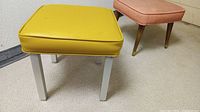 Two mid-century modern footstools on carpeted floor, one with white square legs and yellow vinyl cushion, the other with tapered brown wood legs and orange vinyl cushion.