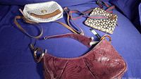 Three designer purses on a purple surface: one burgundy snakeskin handbag, one white patterned crossbody purse with tan accents, and one beige multicolor geometric crossbody purse with blue and red strap.
