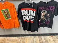 Three t-shirts hanging on hangers against a gray wall and wood floor, clearly showing front graphics and colors: orange Bruno Mars shirt, black RUN DMC shirt, and black Selena Gomez tour shirt.