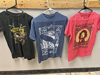 Three rock band T-shirts on hangers against wooden backdrop: Guns N' Roses black, Led Zeppelin blue, Jimi Hendrix red.