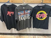 Three black rock band T-shirts hanging on hangers showing logos for The Tubes, The Beatles, and AC/DC