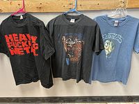 Three rock and roll band T-shirts hanging on hangers against a wooden background, showing front designs for Heavy Fucking Metal, Santana, and Eagles.