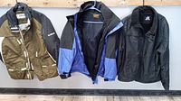 Three ski jackets hanging on a rack: olive green/black Helix, blue/black Gore-Tex, black Zero Exposure