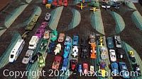 Overview of approximately 40 assorted Hot Wheels vehicles arranged in rows on a carpeted surface, showing cars, semi trucks with trailers, airplanes, tanks, and motorcycles.