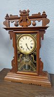 Front right angled view of the antique wooden mantle clock with carved top, glass front panel, and base.