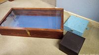 Photo showing a large rectangular wood display box with glass lid, blue interior, and lock, placed next to smaller jewelry boxes in blue and brown.